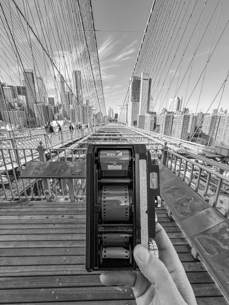 iphone photo of a widelux with the film back off showing what film is loaded in the camera. Brooklyn Bridge as the background.
