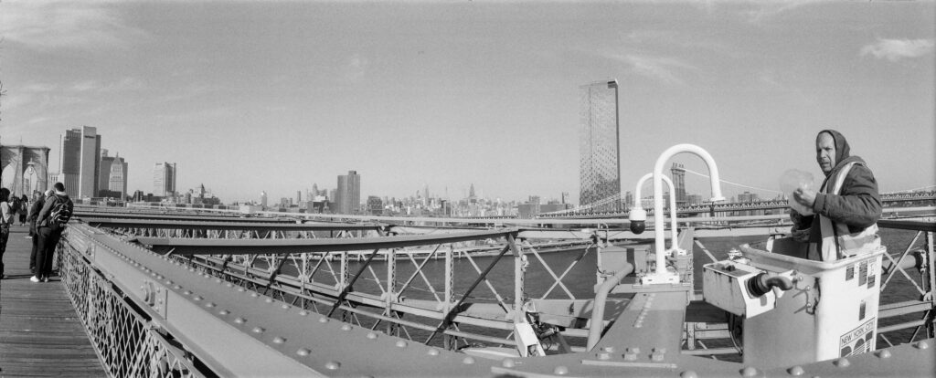 Panoramic black and white photograph of a maintenance worker replacing a security camera on the Brooklyn Bridge with the Manhattan skyline in the background.