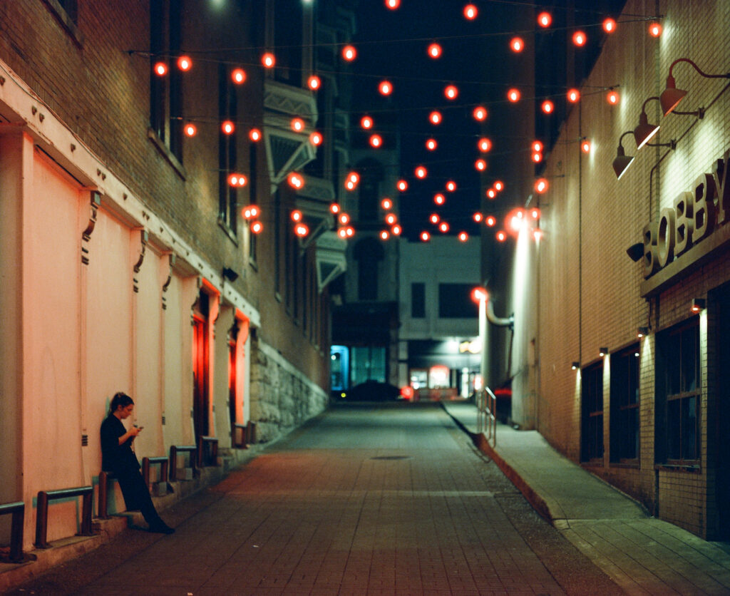 Employee taking a smoke break beneath string lights in Nashville's Printers Alley, photographed at night on a Pentax 67 with CineStill 800T film.