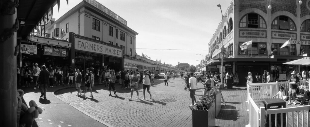 Black-and-white panoramic film photograph of Seattle's Pike Place Market on a busy afternoon, with pedestrians crossing the brick street between historic market buildings. Photographed on a Widelux F7 using Rollei RPX 100 black-and-white 35mm film.