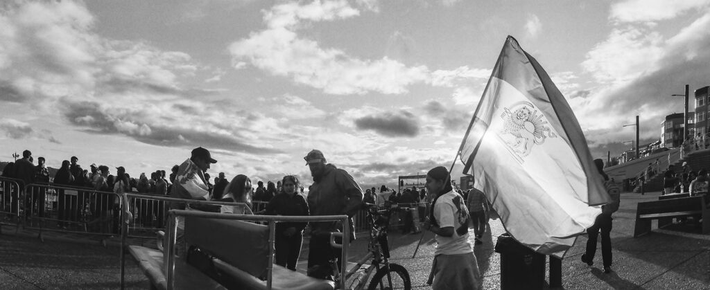 Black-and-white Widelux panoramic photograph of a woman carrying an Iranian Lion and Sun flag at Seattle's FIFA Fan Festival on Pier 62.